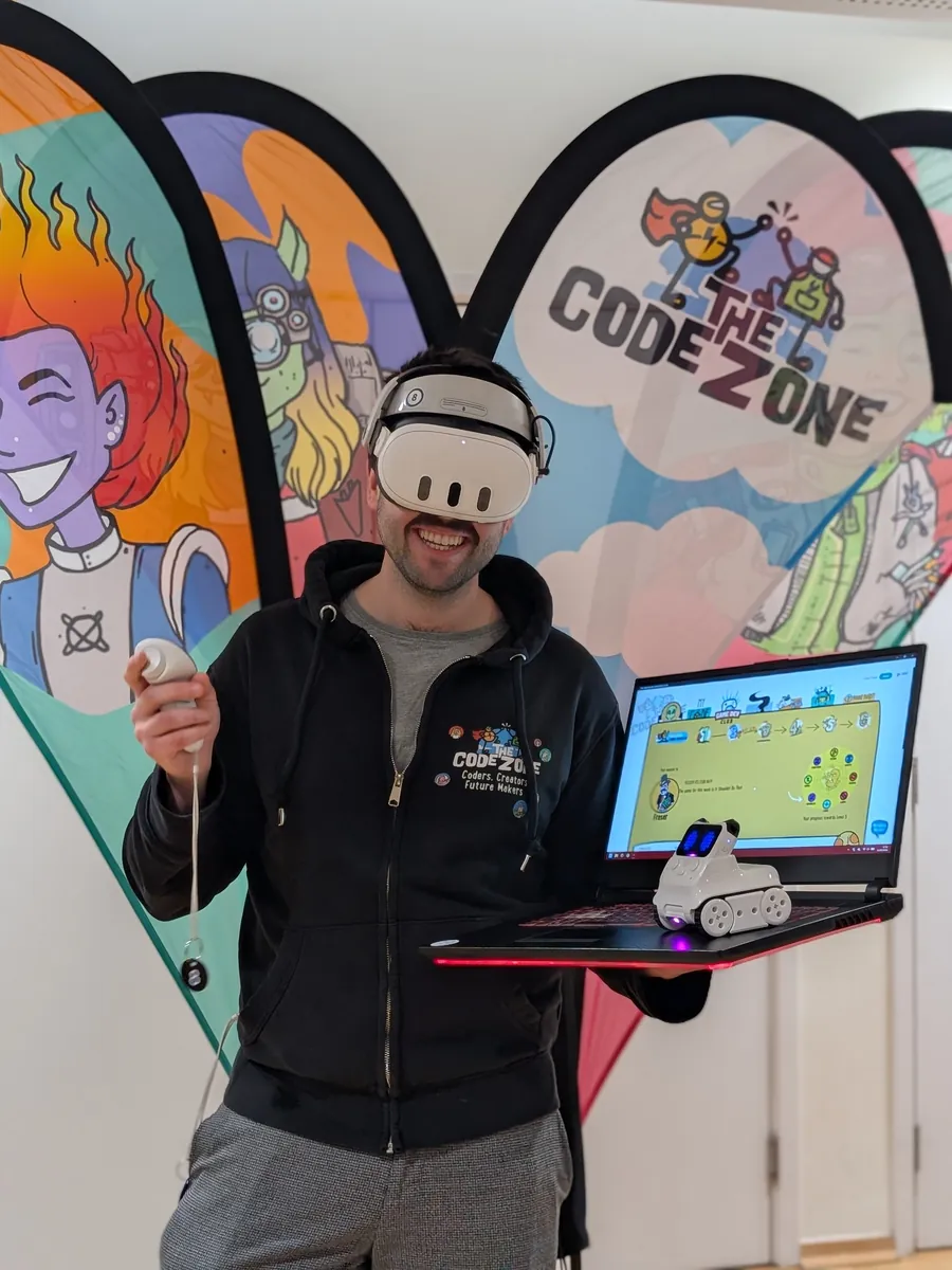 A Code Zone mentor grinning in a virtual reality headset, holding a laptop showing the Game Dev Club map with a small robot sitting on the keyboard, in front of Code Zone banners