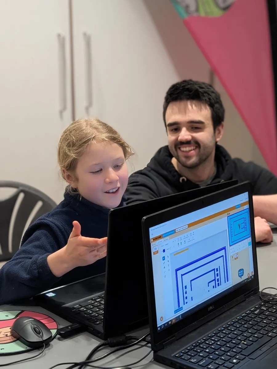 A girl grinning and gesturing at her laptop screen while her mentor sits beside her, smiling at what she has made