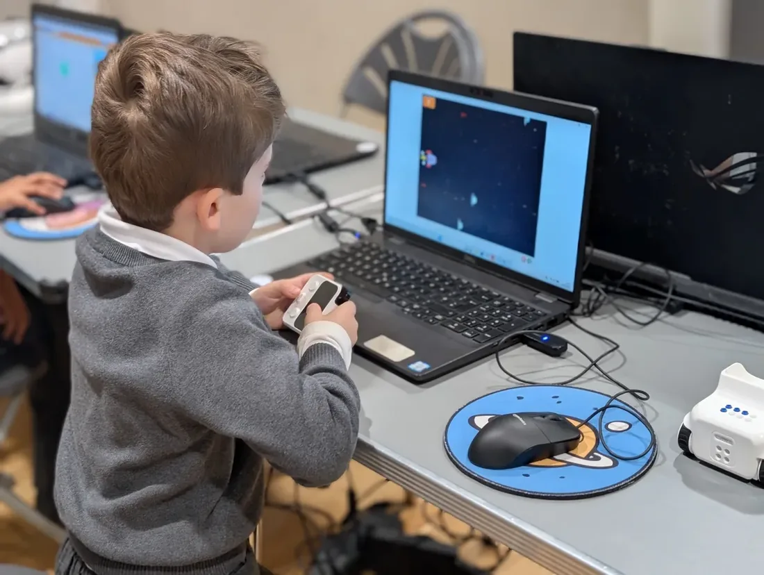 A child at a laptop steering a spaceship game he has built, a small robot on the desk beside him