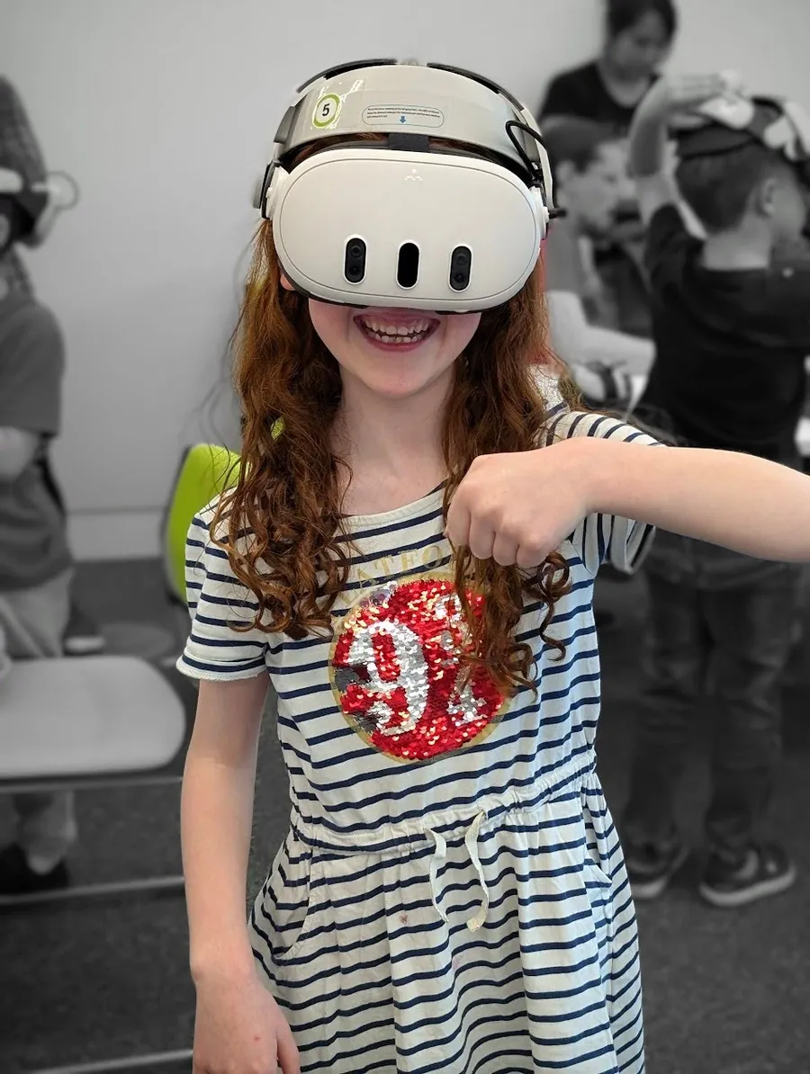 A girl beaming under a virtual reality headset at a club session, with other children playing in the background