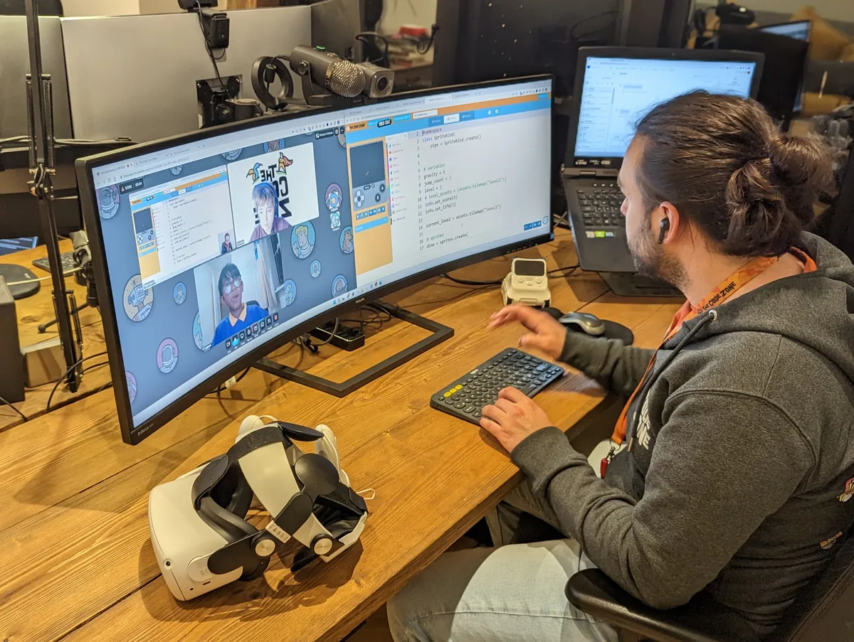 A Code Zone mentor at his desk running a live online club, with members visible in the video call tiles on his monitor beside the game they are building, and a virtual reality headset on the desk.
